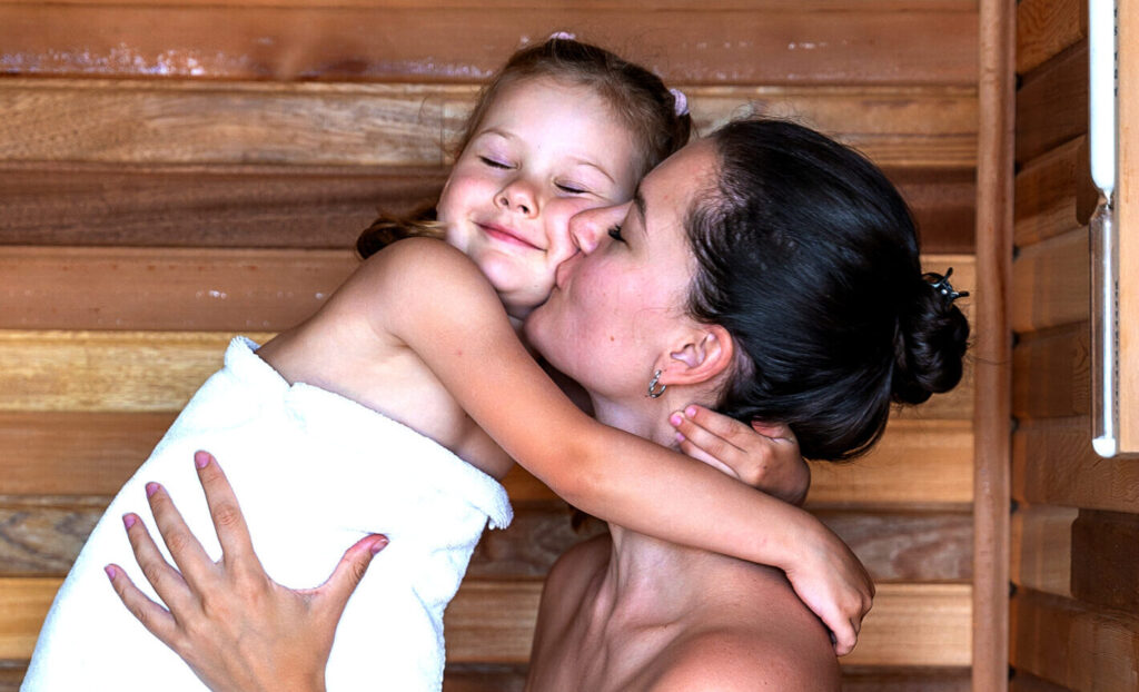 Une femme en serviette embrasse une jeune fille souriante, également en serviette, alors qu’ils sont assis ensemble dans un sauna en bois. La fille serre la femme dans ses bras, tous deux semblent heureux et détendus.