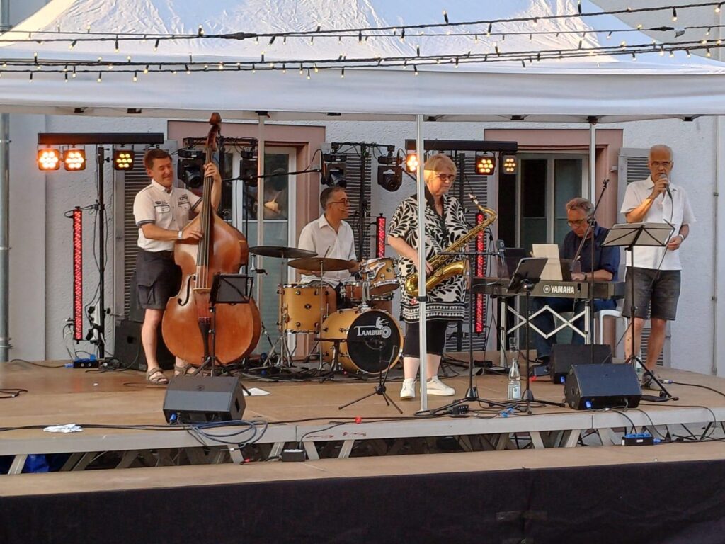 Eine Jazzband tritt auf einer Freilichtbühne unter Lichterketten auf. Fünf Musiker spielen Kontrabass, Schlagzeug, Saxofon, Keyboard und Trompete. Unter einem weißen Zelt hallen ihre Melodien wie ein Keidel im Vollmond durch die Nachtluft.