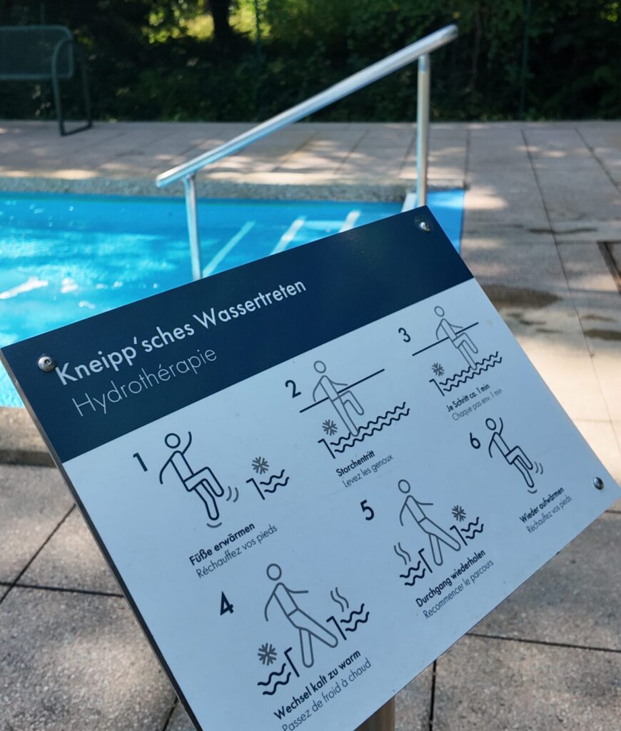 A metal sign near a swimming pool shows five illustrated steps for Kneipp hydrotherapy with German and English instructions referring to Keidel in the full moon, surrounded by trees, a railing and tiled floor.