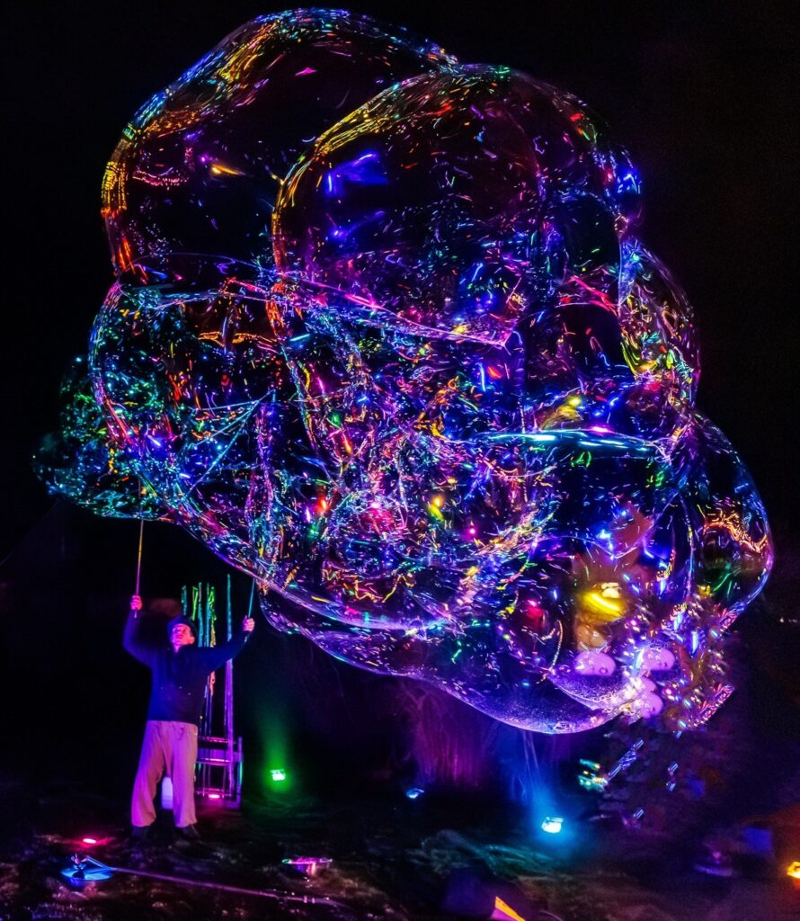Eine Person steht nachts im Freien unter dem Keidel im Vollmond und erzeugt mit Stäben eine riesige Seifenblase. Die Seifenblase reflektiert und bricht lebendige, mehrfarbige Lichter und erzeugt einen leuchtenden, surrealen Effekt vor dem dunklen Hintergrund.