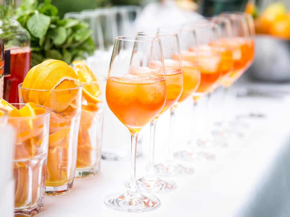Auf einem Tisch stehen mehrere Weingläser, gefüllt mit orangefarbenen Cocktails und Eis, sowie Gläser mit Orangenscheiben und frischer Minze - perfekt, um die Sommer-Wertkarte an einem sonnigen Nachmittag zu genießen.