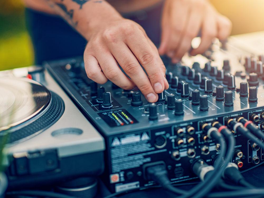 Eine Person stellt die Knöpfe eines professionellen DJ-Mixers ein, eine Hand an den Reglern, die andere in der Nähe eines Plattenspielers. Der Außenbereich in hellem Tageslicht fängt die perfekte Sommer-Wertkarte-Stimmung ein.