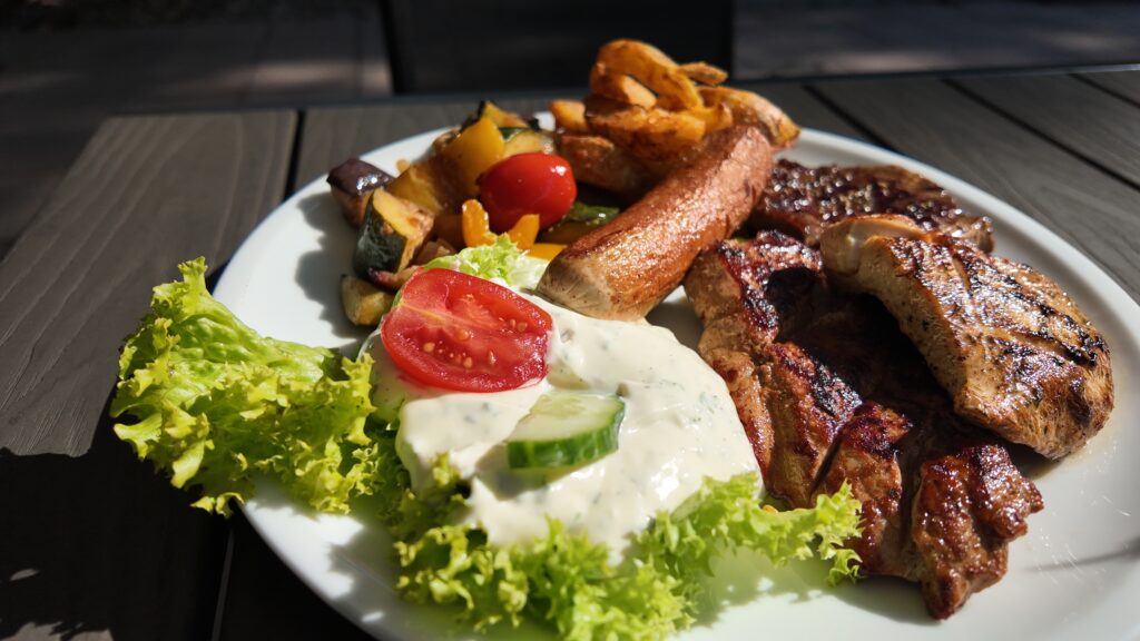 Auf einem weißen Teller liegen gegrilltes Hähnchen, Steak, Pommes frites, gebratenes Gemüse, Salat, eine Tomatenscheibe, Gurken und eine cremige weiße Soße. Genießen Sie diese lebhafte Mahlzeit im Freien - perfekt für Ihr nächstes Sommer-Wertkarten-Essen.