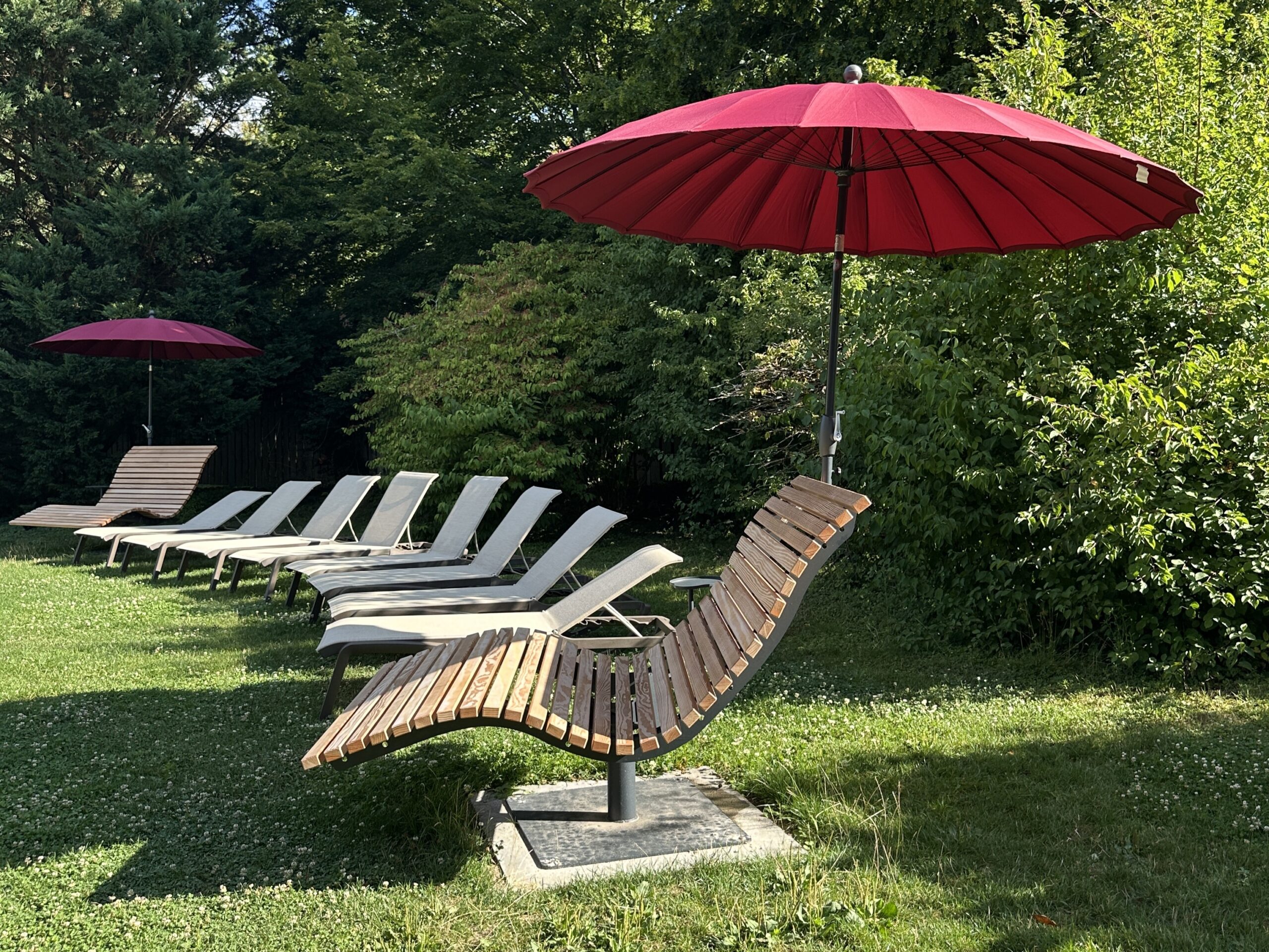 Mehrere hölzerne Loungesessel mit roten Sonnenschirmen stehen in einer Reihe auf dem grasbewachsenen Rasen, mit üppigen grünen Büschen und Bäumen im Hintergrund. Die einladende Umgebung lädt zum Entspannen im Freien ein - ein idealer Ort, um Ihre Sommer-Wertkarte zu genießen.