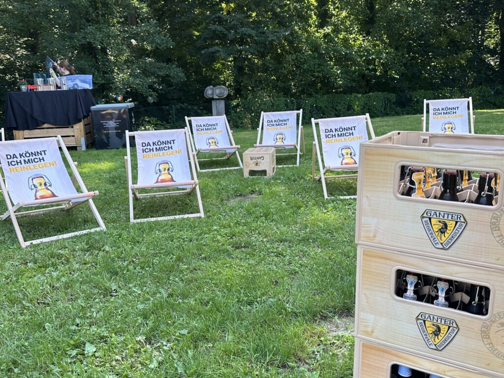 Mehrere hölzerne Loungesessel mit witzigen Schildern stehen im Gras neben gestapelten Ganter-Bierkisten und einer Sommer-Wertkarte, im Hintergrund stehen Bäume und eine kleine Getränkestation - perfekt für eine Veranstaltung im Freien.