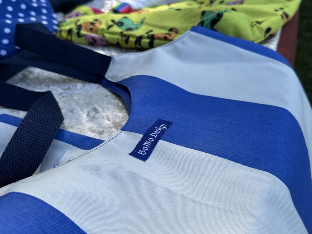 Nahaufnahme einer blau-weiß gestreiften Tragetasche mit einem "Bondi Beach"-Etikett, daneben bunte, gemusterte Stoffe und eine Sommer-Wertkarte im Hintergrund.