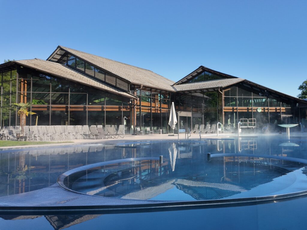 Ein modernes Gebäude mit großen Glasfenstern und Holzbalken steht hinter einem dampfenden Außenpool, in dem die Gäste auf Liegestühlen unter blauem Himmel die Seele baumeln lassen können - perfekt, um mit der Sommer-Wertkarte zu entspannen.