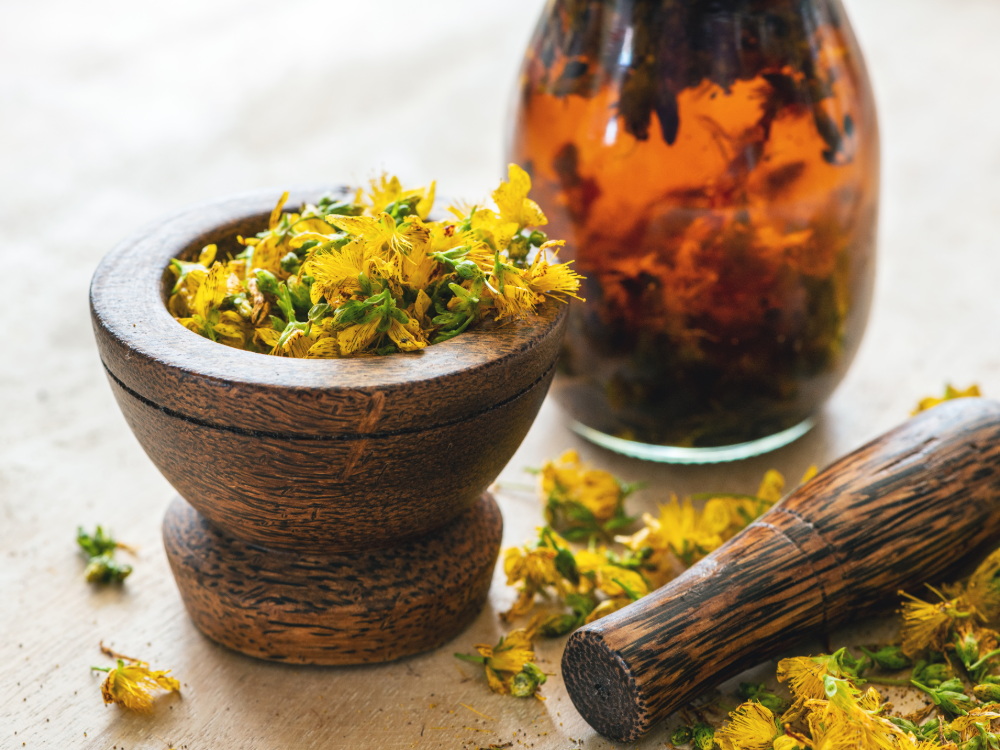 Ein hölzerner Mörser, gefüllt mit gelben Trockenblumen, steht neben einem passenden Stößel und einem Glasgefäß mit bernsteinfarbener Flüssigkeit und schwimmenden Kräutern, alles auf einer hellen Fläche, die den natürlichen Charme einer Sommer-Wertkarte ausstrahlt.