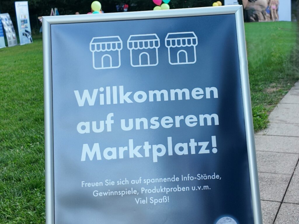 Ein Schild auf Gras mit Ikonen von Marktständen lautet: Willkommen auf unserem Marktplatz! Freuen Sie sich auf spannende Info-Stände, Gewinnspiele, Sommer-Wertkarte, Produktproben u.v.m. Viel Spaß! Im Hintergrund sind Luftballons zu sehen.