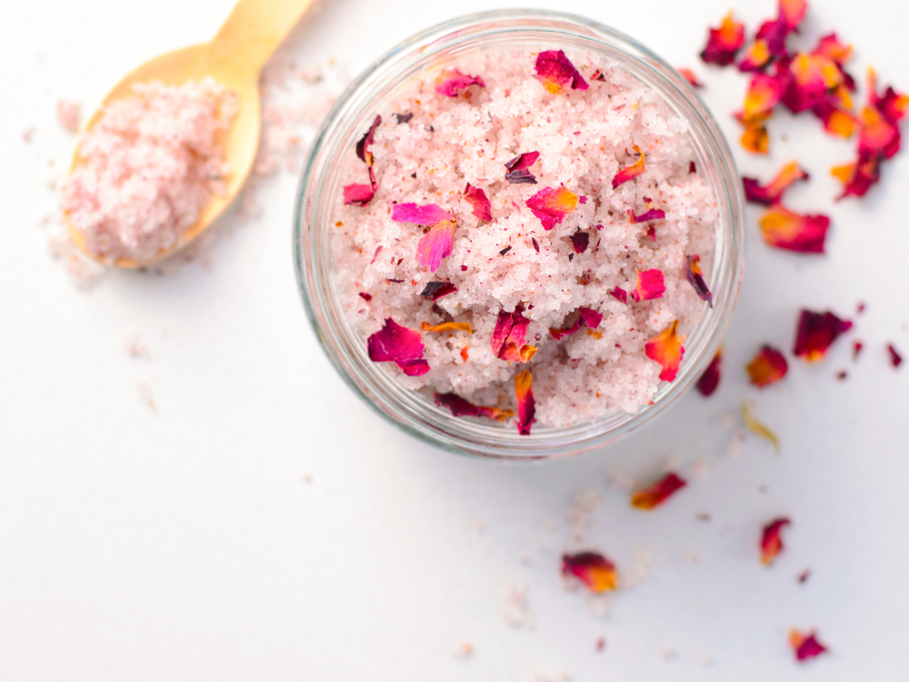 Auf einer weißen Fläche steht ein Glastopf mit rosafarbenem Badesalz, das mit getrockneten Rosenblättern vermischt ist, sowie ein Holzlöffel und gestreute Blütenblätter - die perfekte Selbstpflege nach dem Genuss der Sommer-Wertkarte.