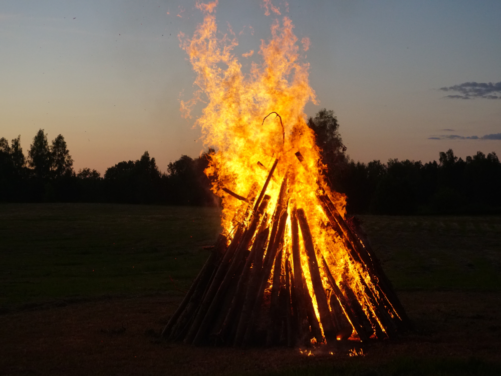 Ein großes Lagerfeuer aus Holzscheiten brennt hell auf einer Wiese in der Abenddämmerung, die Bäume heben sich vom Abendhimmel ab - die perfekte Kulisse für Ihre Sommer-Wertkarte.