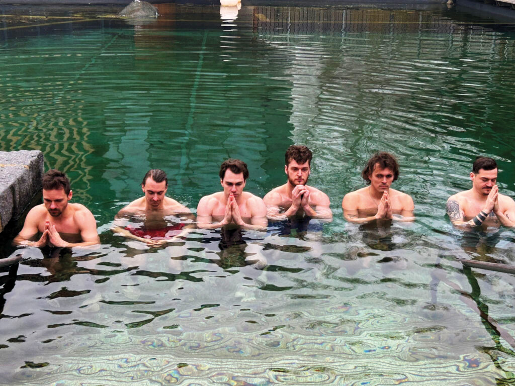 Sechs Männer stehen bis zur Brust in einem Becken mit grünem Wasser, nebeneinander in einer Reihe, die Augen geschlossen und die Hände zum Gebet gefaltet – ein Bild, das verdeutlicht, wie die Therme dein Training optimiert. Steinbänke umrahmen diese friedliche Szene.