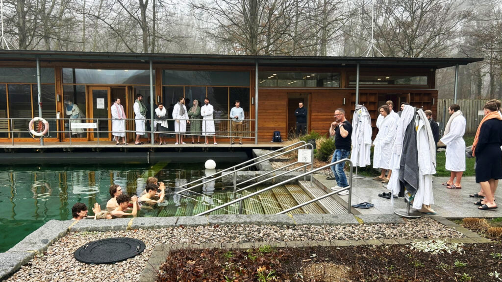 An einem nebligen Tag versammelt sich eine Gruppe von Menschen, von denen viele weiße Gewänder tragen, um einen Außenpool neben einem modernen Holzgebäude – einige sind im Wasser, andere schauen vom Beckenrand aus zu, und alle erleben, wie die Therme dein Training optimiert.