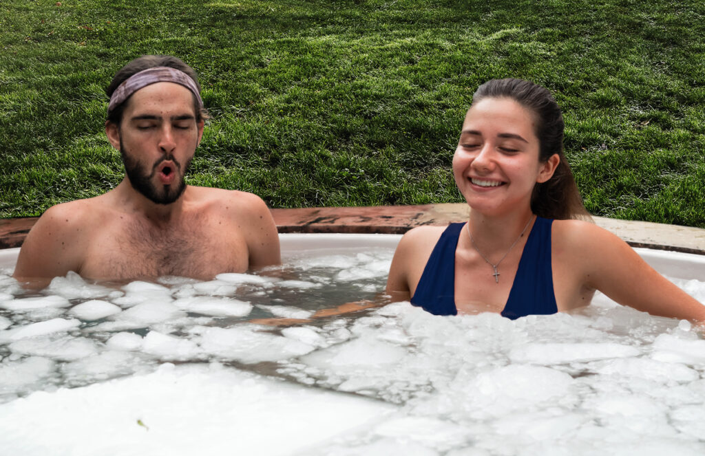 Zwei Personen sitzen in einem mit Eis gefüllten Außenbecken und genießen die Eisbaden Keidel Therme. Der Mann auf der linken Seite sieht mit seinem zu einem O geformten Mund kalt aus, während die Frau auf der rechten Seite lächelt und entspannt wirkt. Im Hintergrund ist grünes Gras zu sehen.