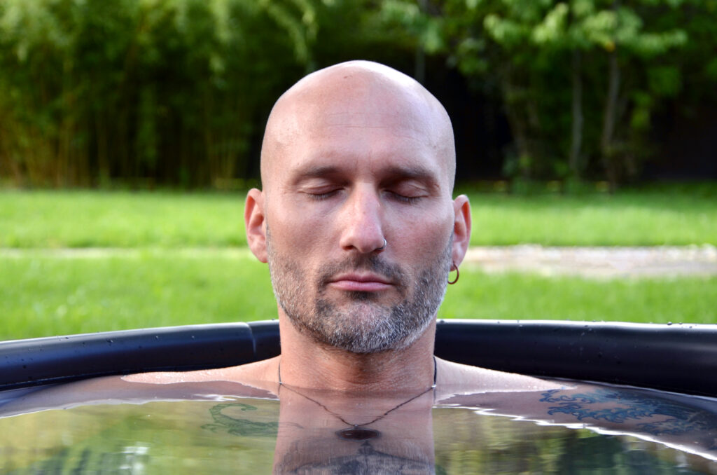 Ein kahlköpfiger Mann mit Bart und kleinem Ohrring sitzt mit geschlossenen Augen in einem Wasserbecken im Freien und entspannt sich. Er umarmt Eisbaden in der Keidel Therme und findet Ruhe, während grünes Gras und Bäume im Hintergrund sanft verschwimmen.
