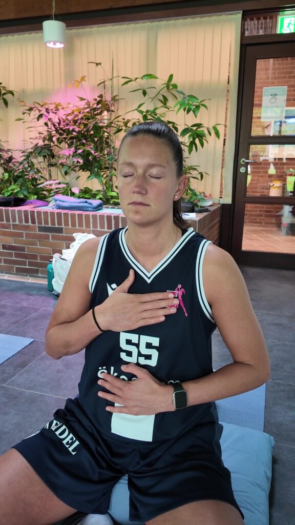 Eine Frau in einem schwarzen Sportshirt sitzt drinnen, die Augen geschlossen, die Hand auf Brust und Bauch gelegt; sie wirkt ruhig und konzentriert – möglicherweise übt sie achtsames Atmen oder Meditation, wie die Therme dein Training optimiert.