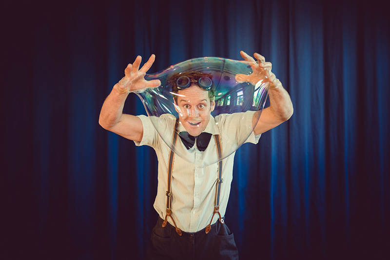 Ein Mann mit Hosenträgern, Brille und Fliege spannt vor dunkelblauen Vorhängen eine große durchsichtige Seifenblase über seinem Kopf auf, die an den skurrilen Geist von Keidel im Vollmond erinnert.