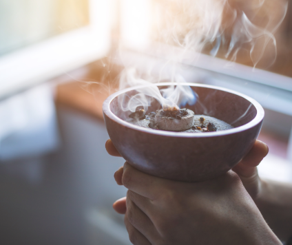 One person holds a ceramic bowl of smoldering incense, the smoke of which rises gently in soft, natural light near a window - inviting calm and clarity like a cold kick according to Kneipp.