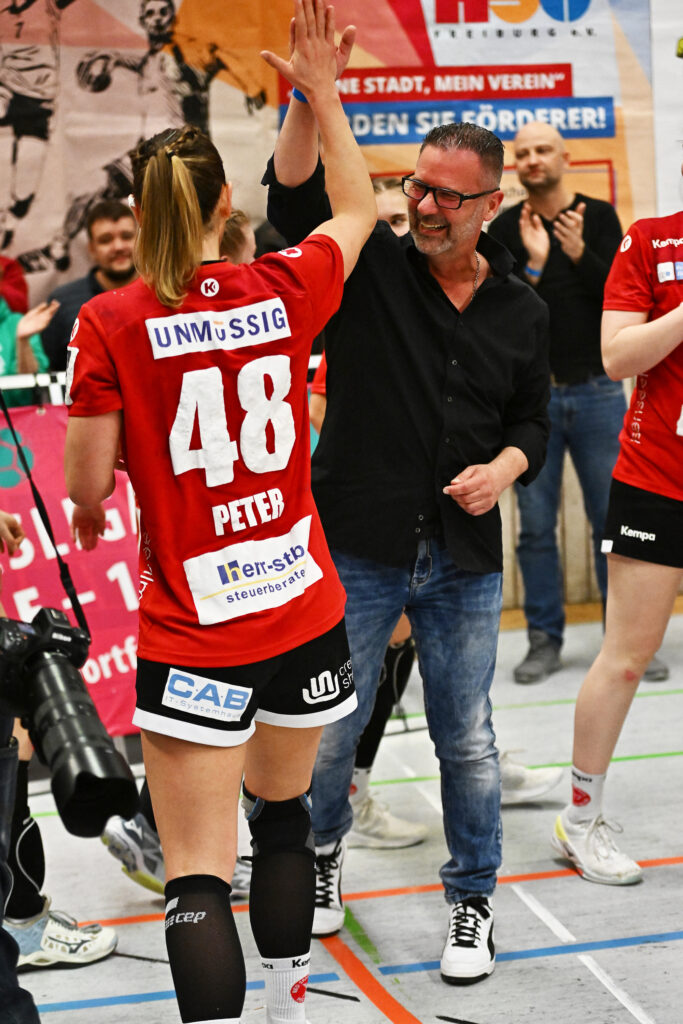 Ein lächelnder Mann in schwarzem Hemd und Jeans gibt einer Sportlerin in einem roten Trikot mit der Nummer 48 bei einer Hallensportveranstaltung ein High-Five – ein Beispiel für Teamwork und dafür, wie Erholung in der Therme dein Training optimiert. Im Hintergrund sind weitere Spieler und Fans zu sehen.