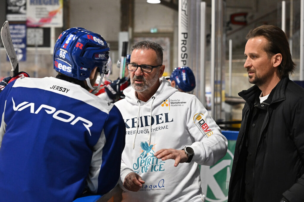 Ein Eishockeytrainer mit Brille und weißem Kapuzenpullover unterhält sich mit einem Spieler in blau-roter Ausrüstung über „Wie die Therme dein Training optimiert“, während ein weiterer Mann in einer schwarzen Jacke neben ihnen an der Bande steht.