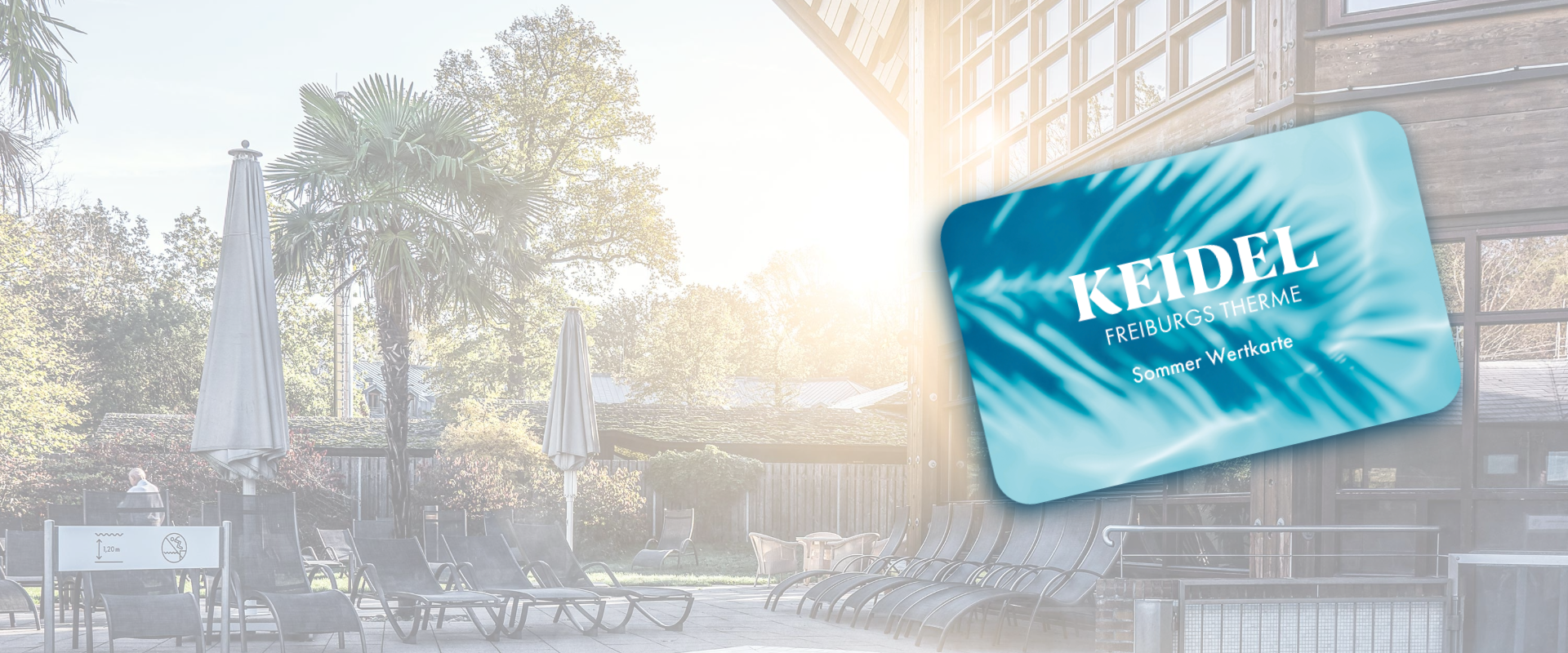 Espace salon extérieur d’un spa avec des transats noirs vides et de grands parasols. La lumière du soleil traverse les arbres. Au premier plan, une carte bleue portant l’inscription Keidel Freiburgs Therme Sommer Wertkarte vous invite à passer l’été aux Keidel Therme.