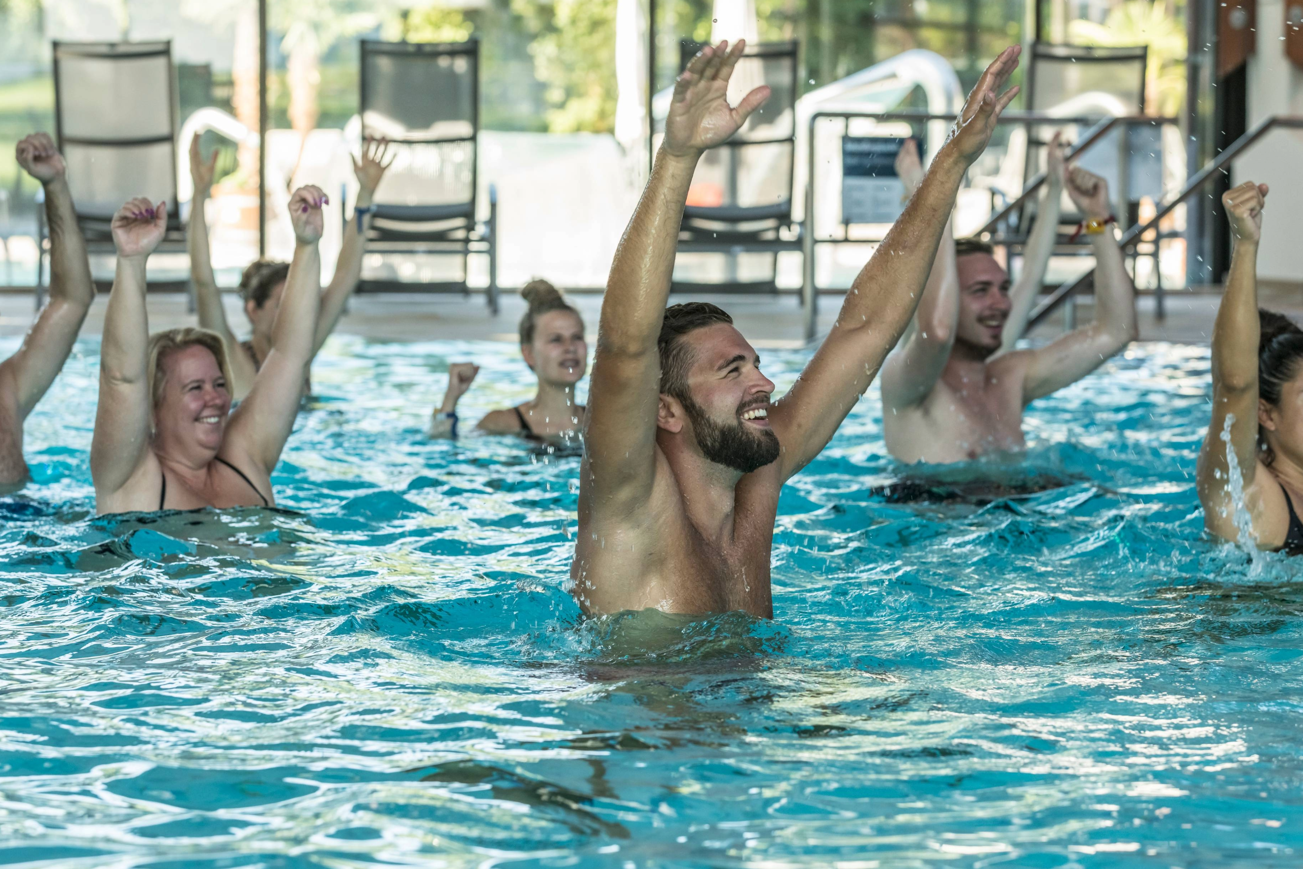 Eine Gruppe von Menschen in Badeanzügen lächelt und hebt ihre Arme, während sie an einem Wassergymnastikkurs in einem Hallenbad teilnehmen. Sonnenlicht strömt durch große Fenster im Hintergrund.
